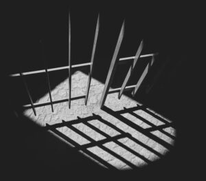 A dark black-and-white photo of prison bars casting a shadow on a stone floor.